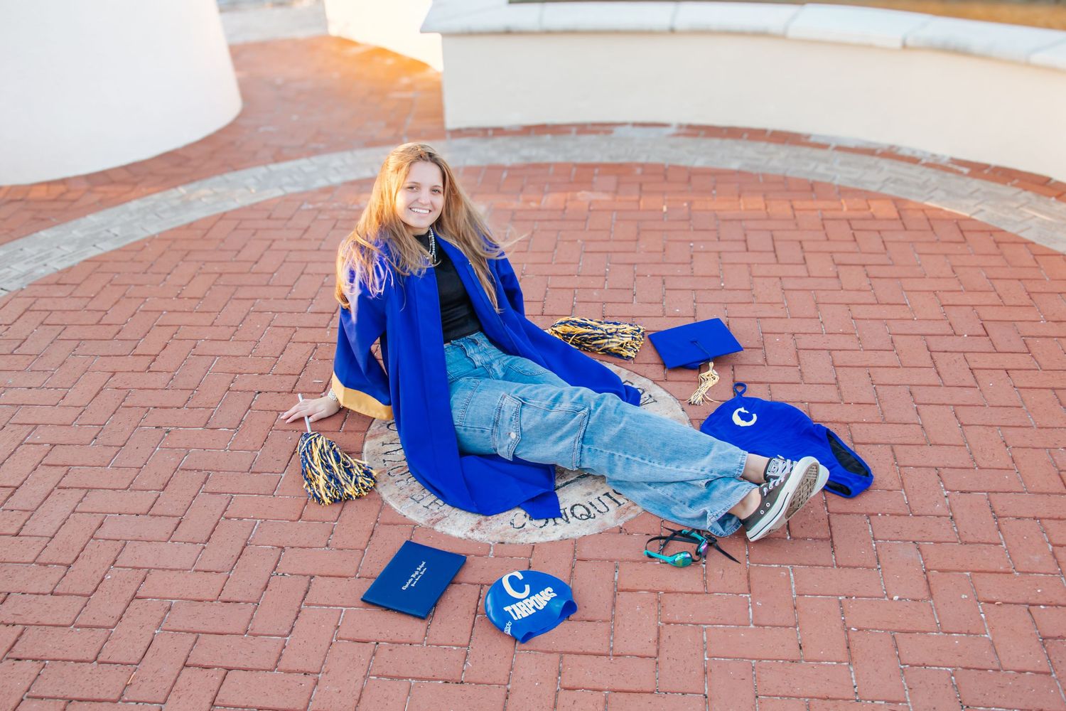 Young woman in blue graduation gown and cap sitting on brick plaza with diploma, celebrating her graduation day.