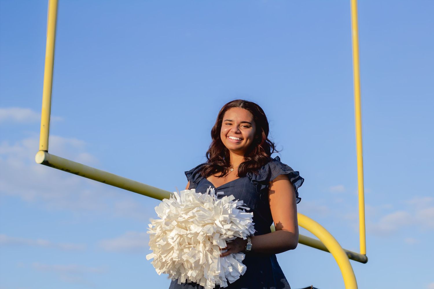 Cheerleader holding white pom-poms stands in front of yellow football goal post against blue sky.
