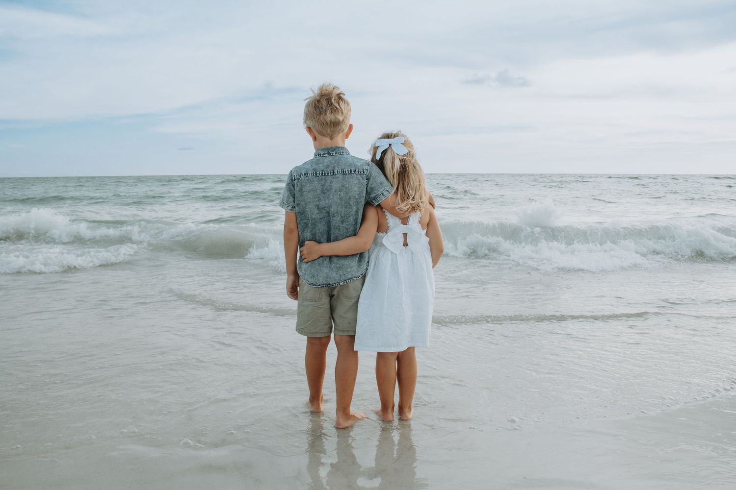 Two children standing barefoot on a sandy beach shore, looking out at ocean waves together.
