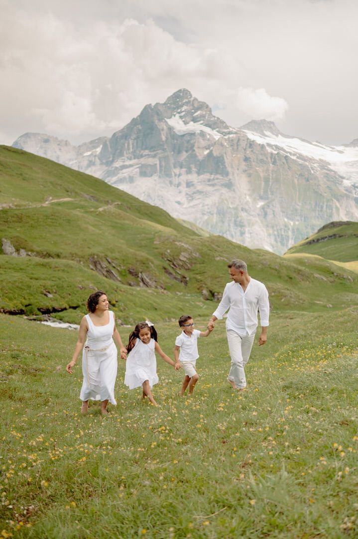 A family in white outfits walks up a grassy mountainside with snow-capped alpine peaks in the background.