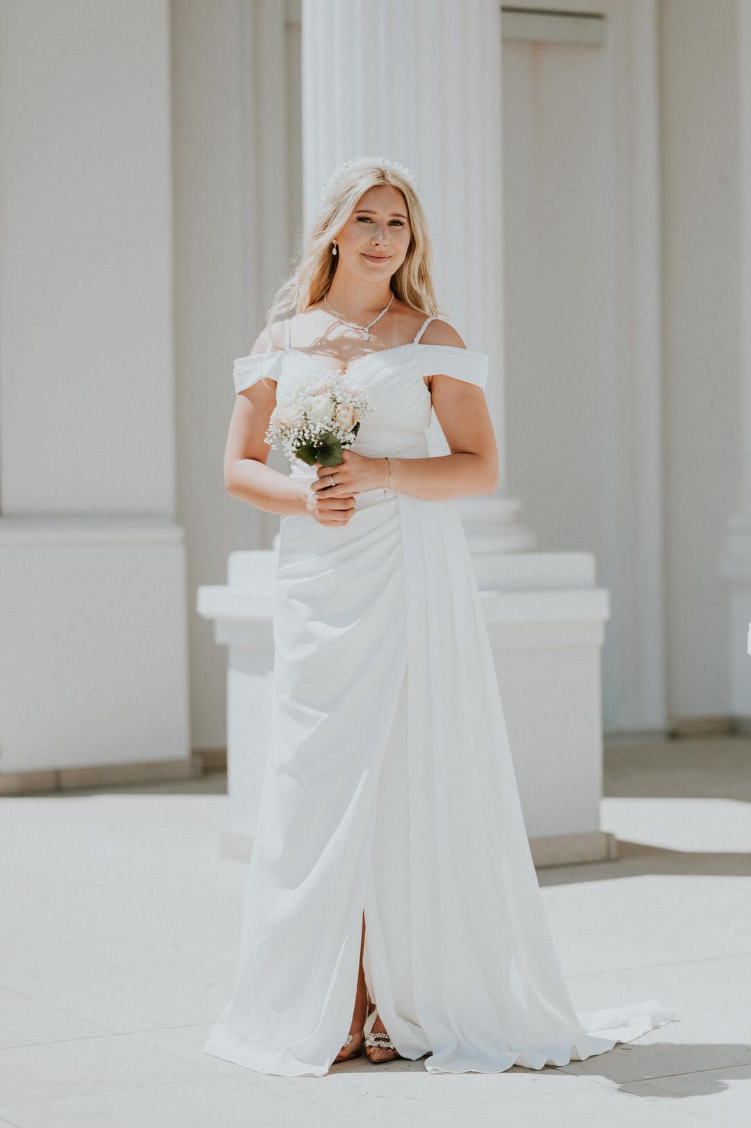 Elegant bride in off-shoulder white wedding gown holds small bouquet against white column backdrop.