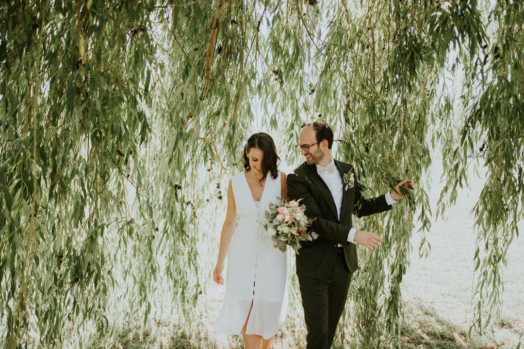 Elegant couple in wedding attire posed beneath weeping willow tree.