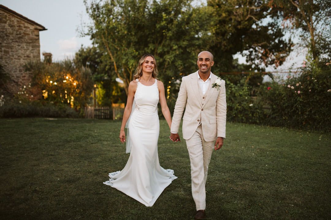 Couple in wedding attire walking together on garden lawn at sunset.