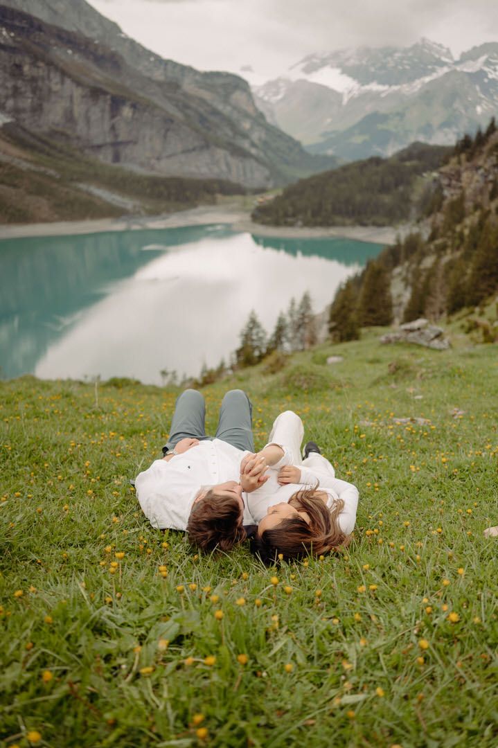 Two people lie in a grassy meadow overlooking a turquoise alpine lake surrounded by majestic mountain peaks.