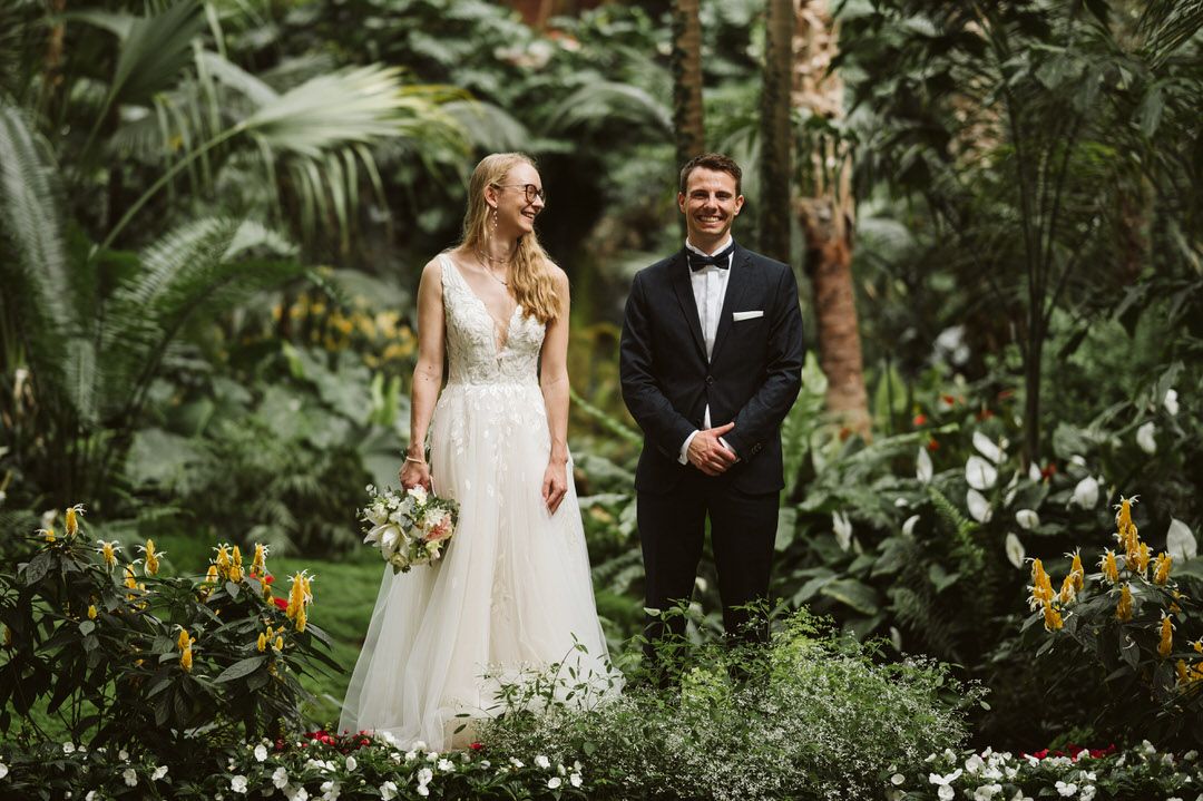 Wedding couple standing in lush tropical garden surrounded by exotic plants and flowers.
