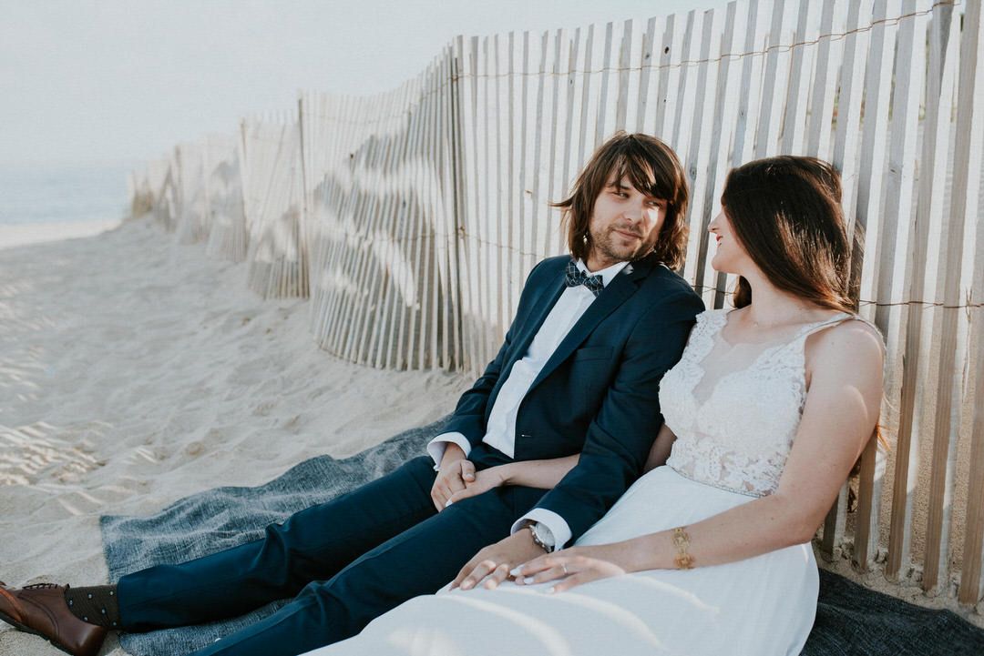 Relaxed wedding couple sitting by beach fence during an intimate coastal photoshoot.