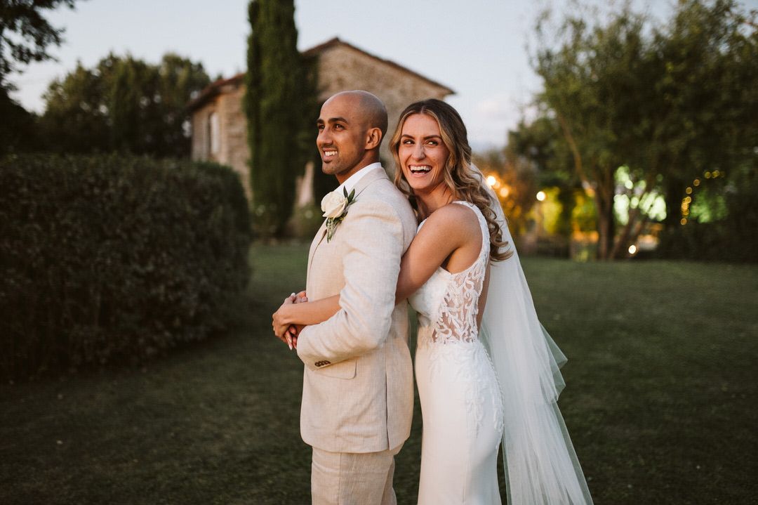 Wedding couple in light-colored attire pose back-to-back in a garden setting during golden hour.