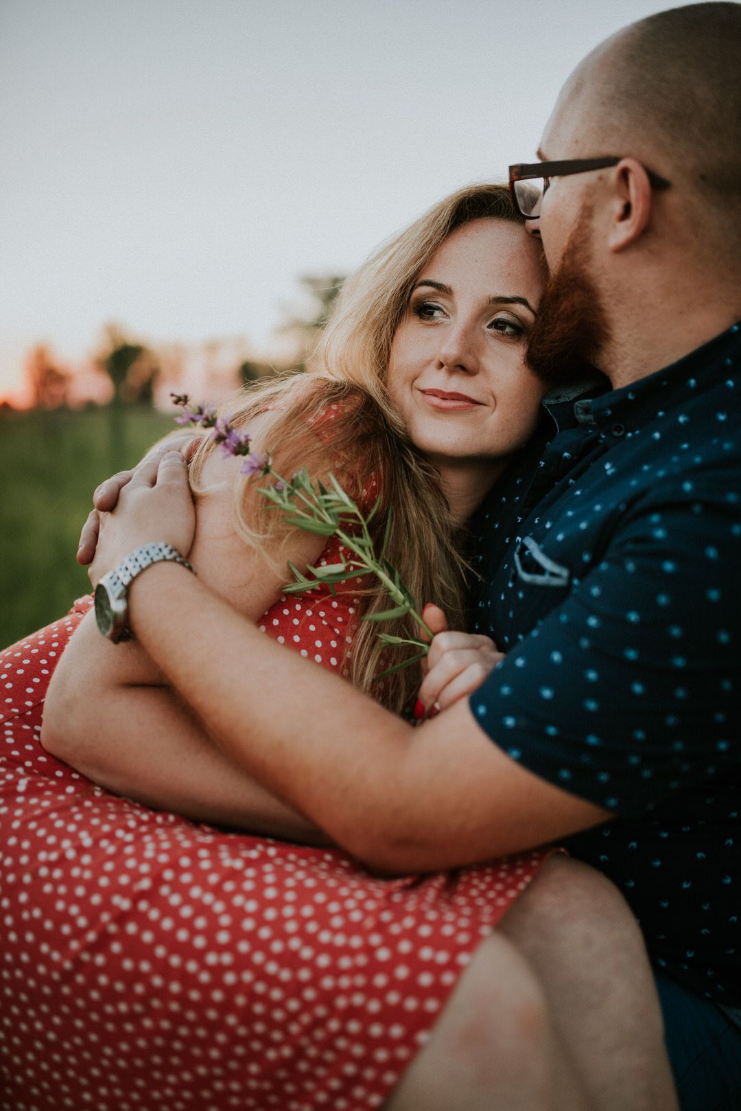 Romantic couple embracing outdoors with red polka dot dress and navy shirt in warm sunlight.