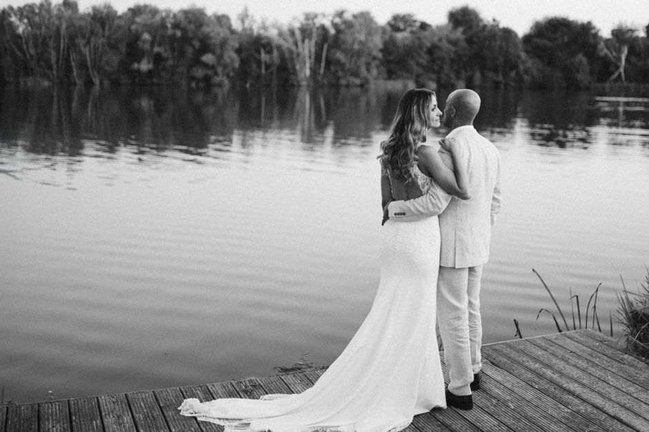 Newlyweds embrace on lakeside dock with flowing white dress trailing behind in monochrome.