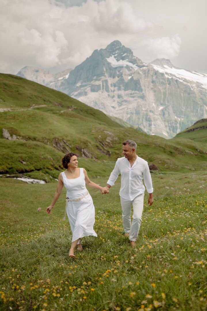 Couple in white outfits walks together through an alpine meadow with dramatic mountain peaks in the background.