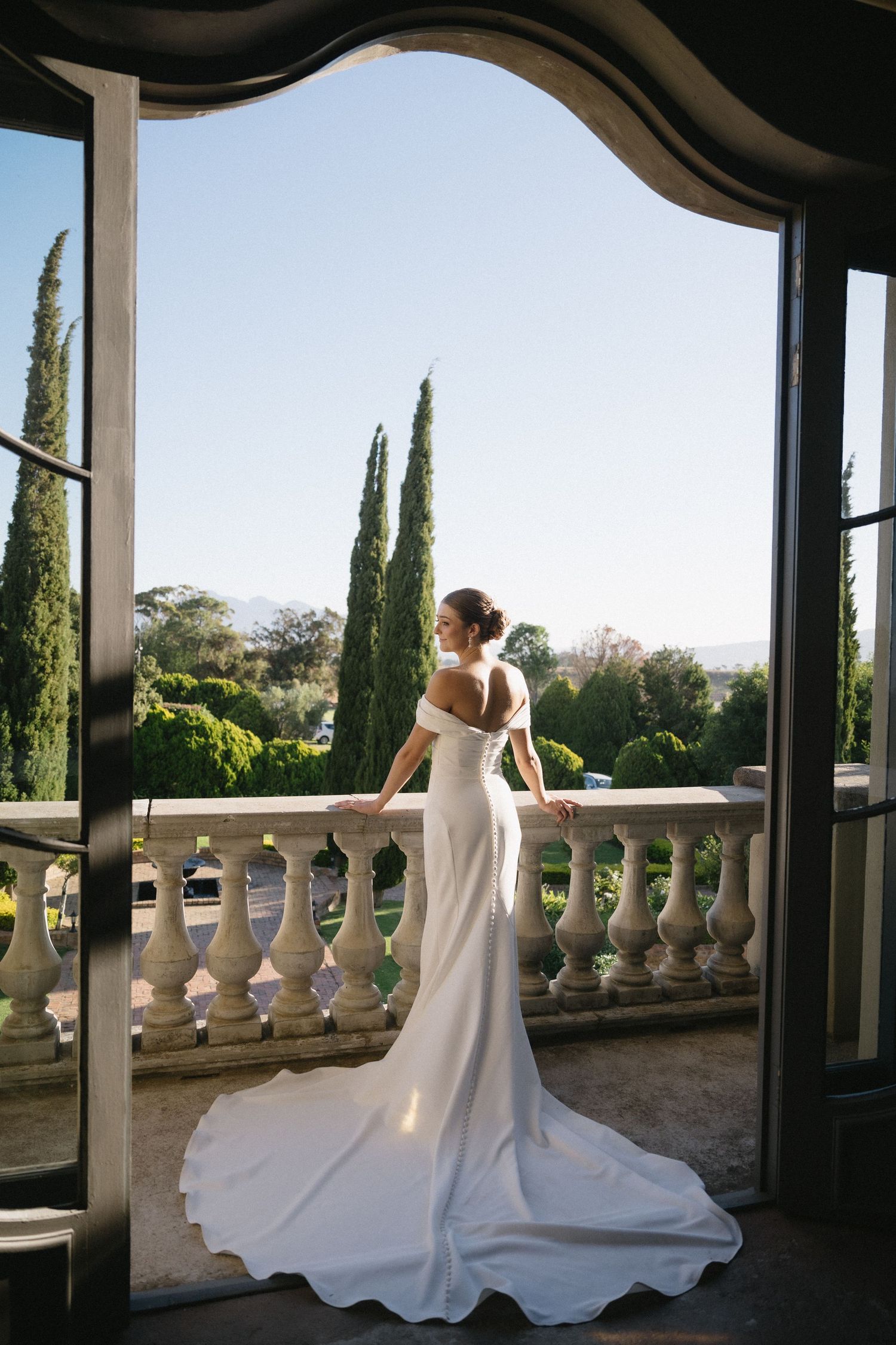 Elegant bride in flowing white gown stands on ornate balcony with cypress trees and garden views in background.