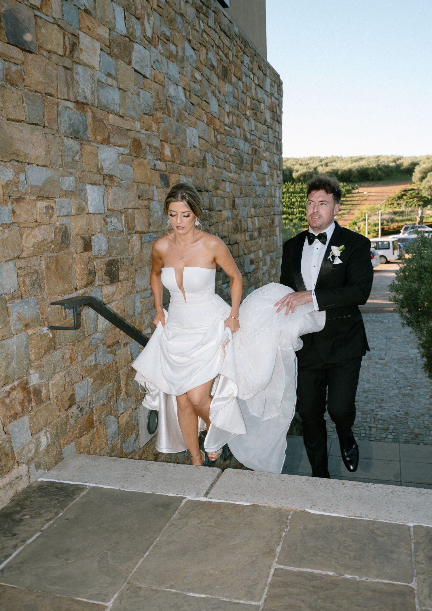 A couple at a stone building venue with bride lifting her white dress while walking up stone steps in casual wedding moment.