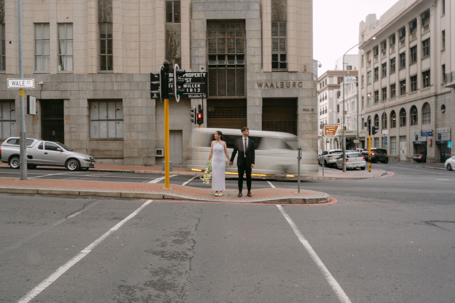 People with umbrellas wait at a crosswalk on a rainy day in an urban downtown intersection.