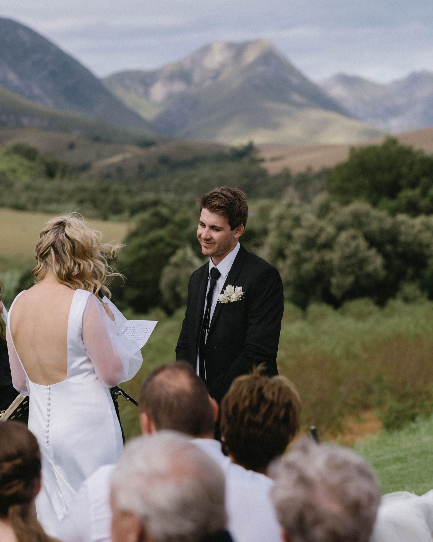 Outdoor wedding ceremony with mountain backdrop as couple exchanges vows in front of seated guests.