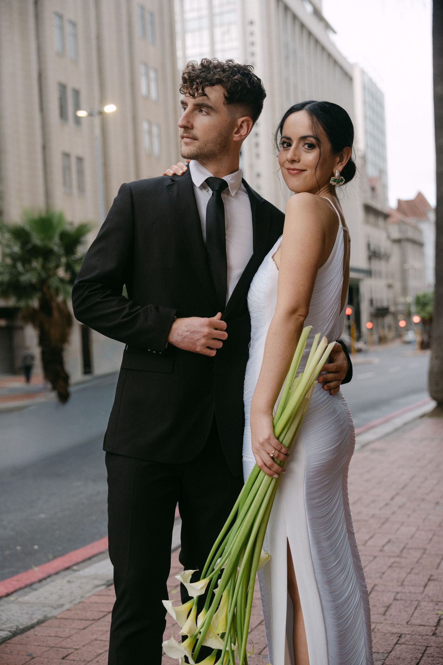Romantic couple in wedding attire share intimate moments while walking along a city street.