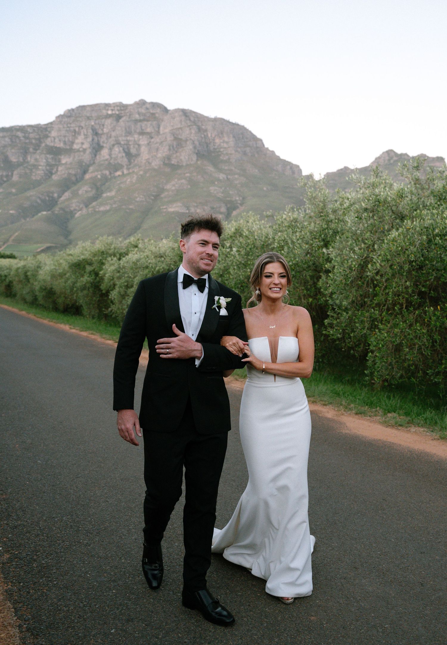 A wedding couple walks along a road with majestic mountain scenery and green foliage in the background.