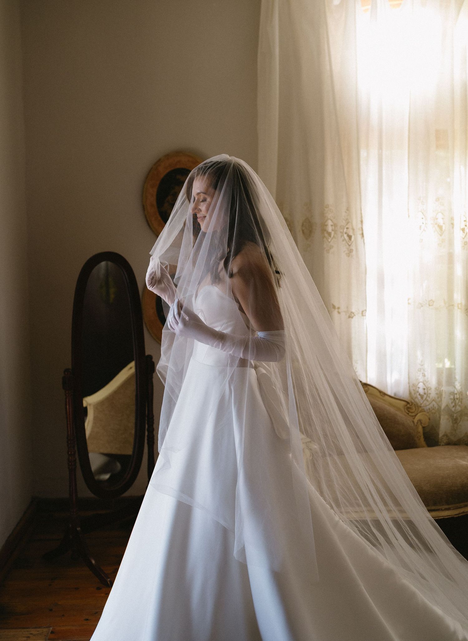 A bride in a flowing white gown and veil stands elegantly by a vintage mirror.
