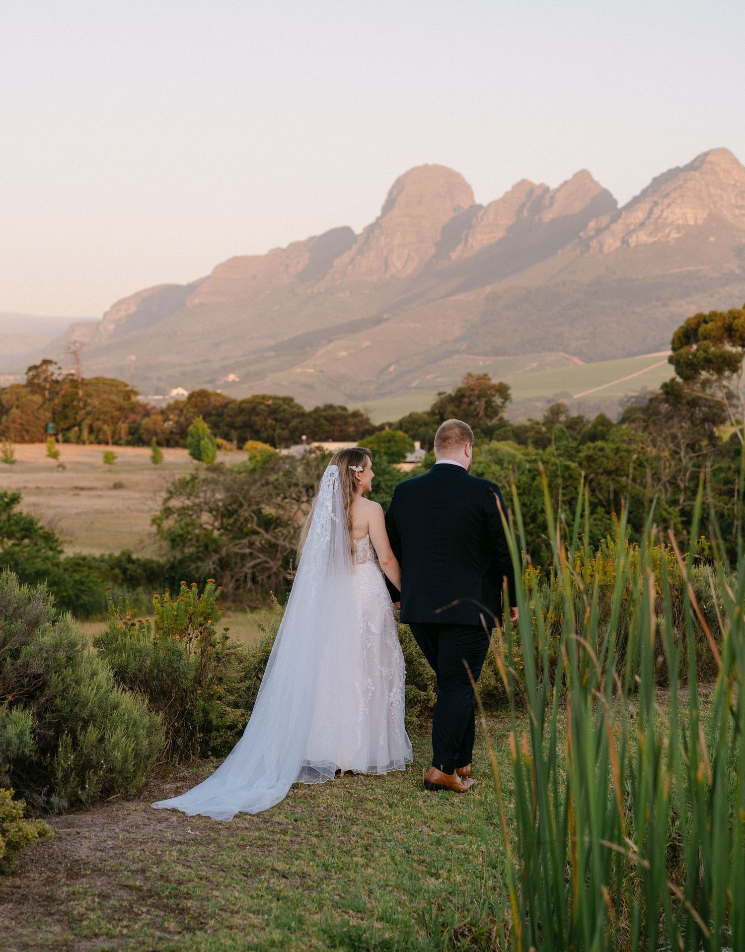 A wedding couple stands overlooking scenic mountain landscape at sunset.