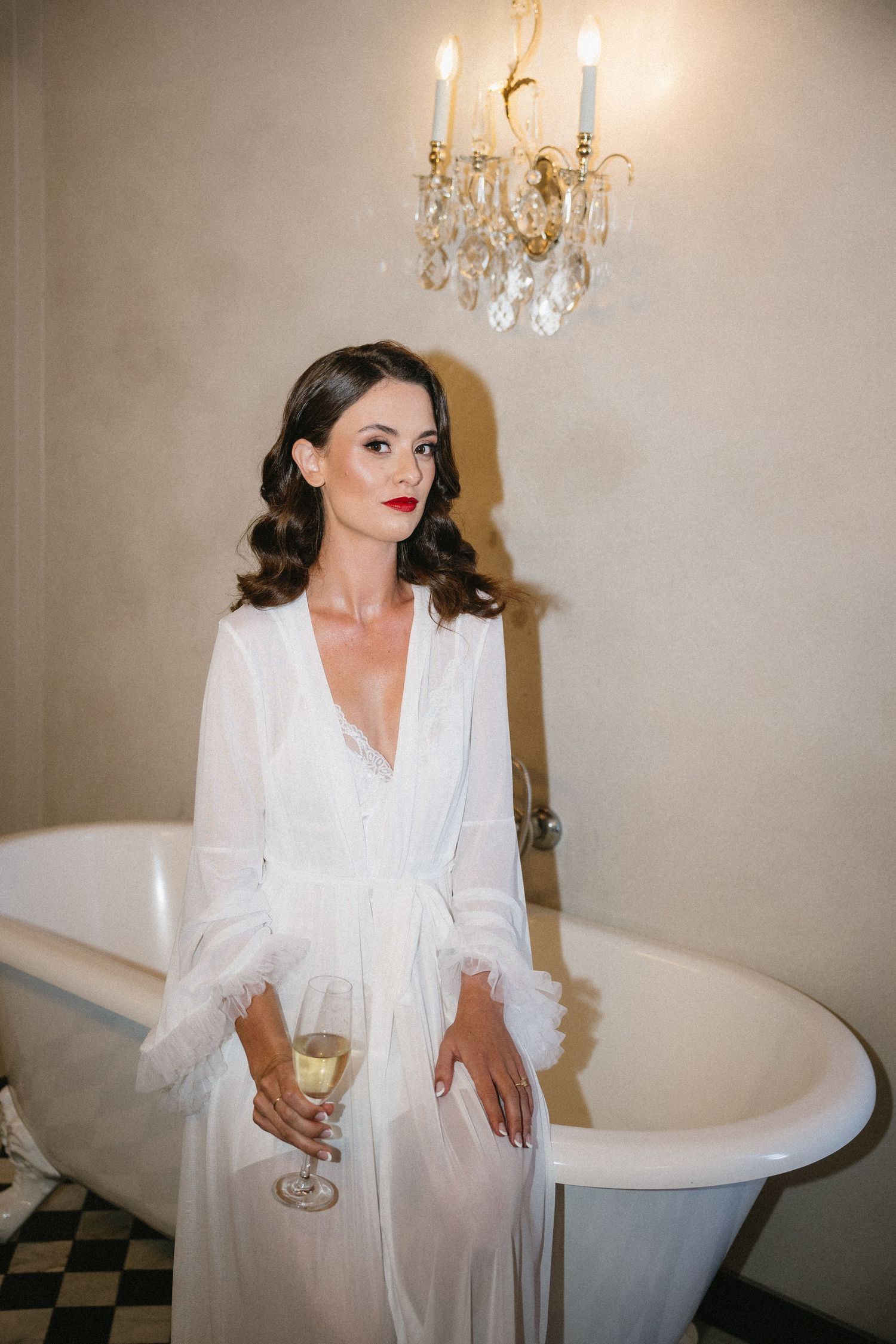 Elegant woman in white silk robe holds champagne glass beside vintage clawfoot tub with crystal chandelier above.