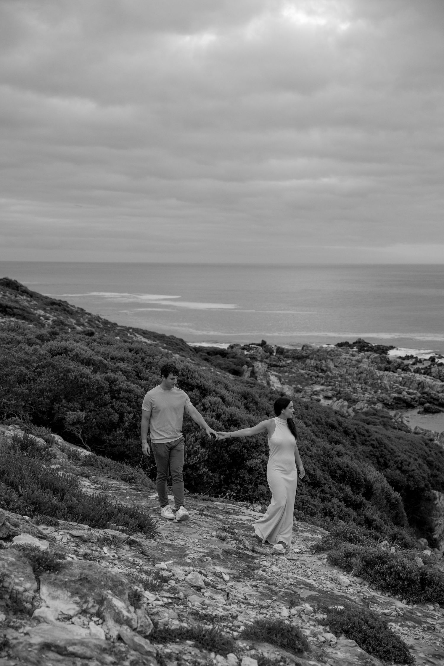 A couple holds hands while walking along a scenic coastal trail with dramatic ocean views and cloudy skies above.