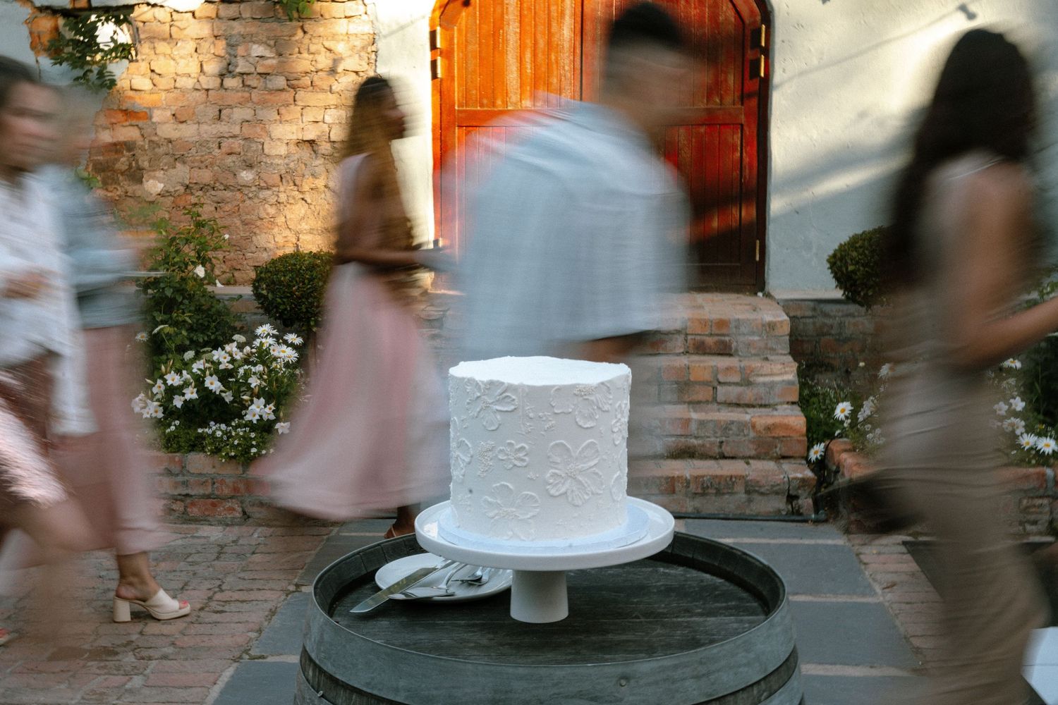 White wedding cake on stand with blurred motion of guests walking past.