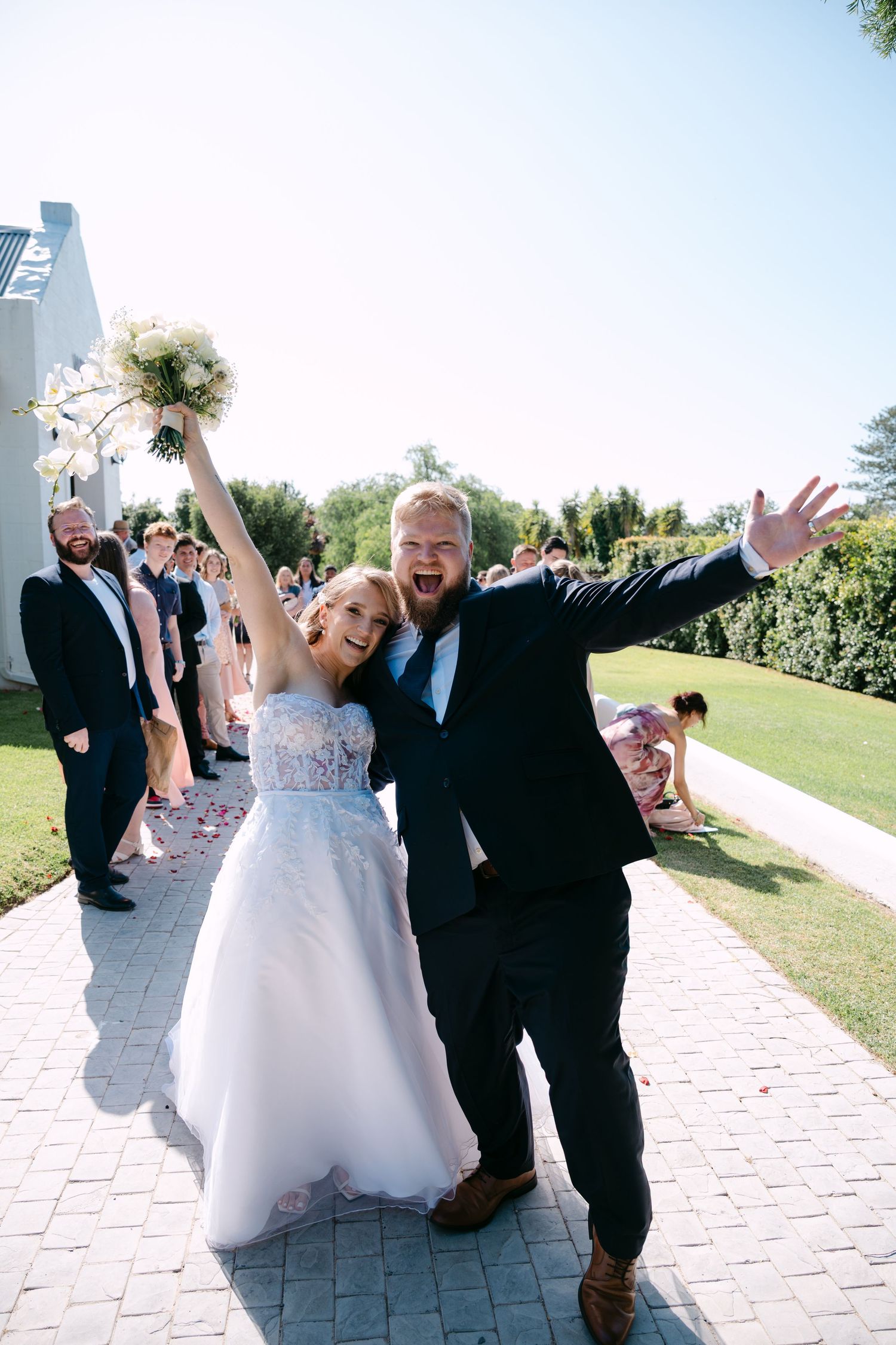 Newlyweds celebrating joyfully on a stone pathway outside a white building on their wedding day.