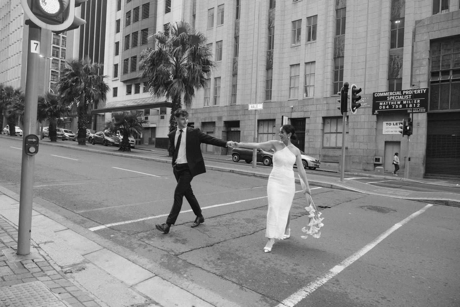 Pedestrians cross a city intersection while a person in white clothing walks along the sidewalk in black and white.