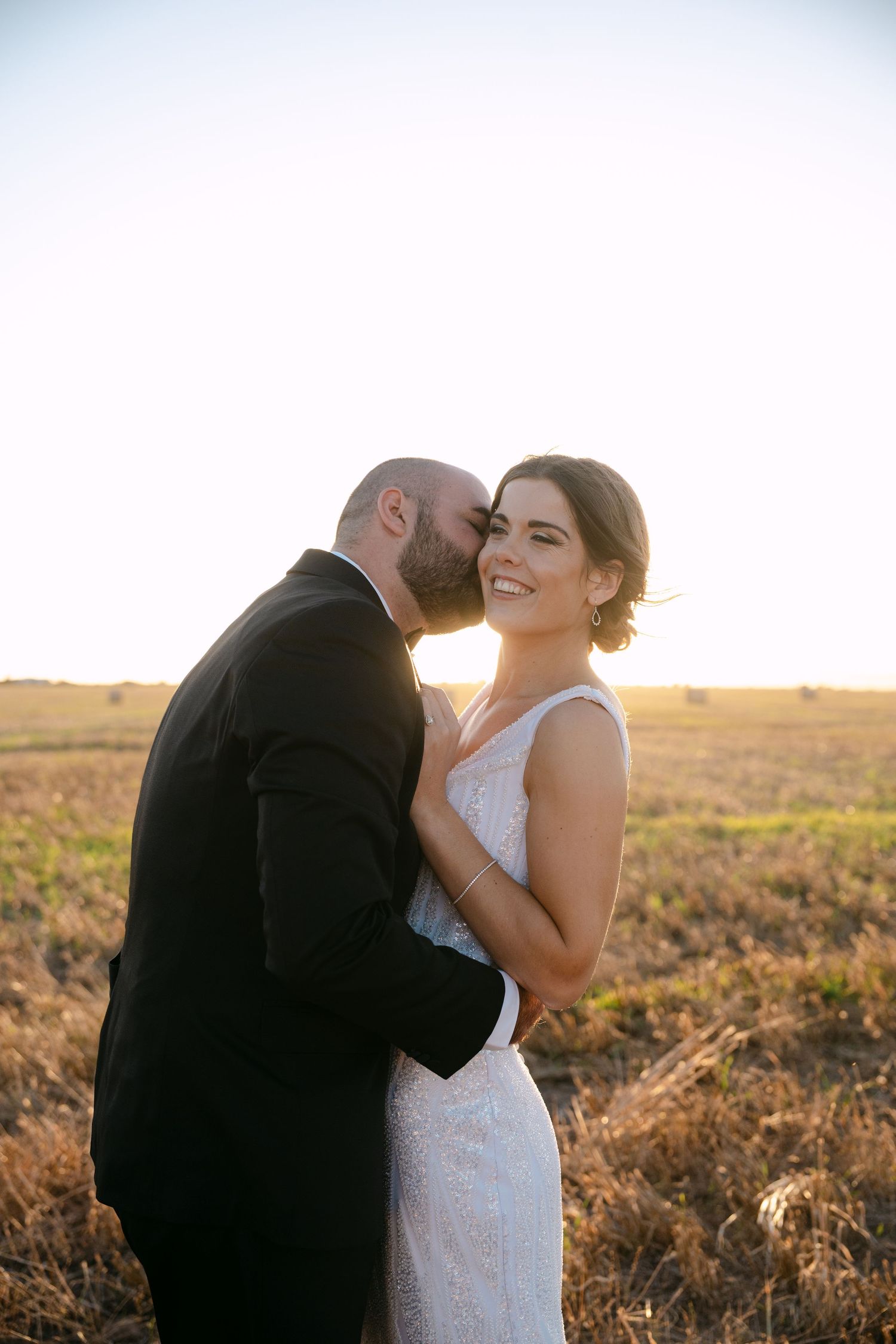 Newlyweds share an intimate moment in a field at sunset, with golden light illuminating their wedding attire.