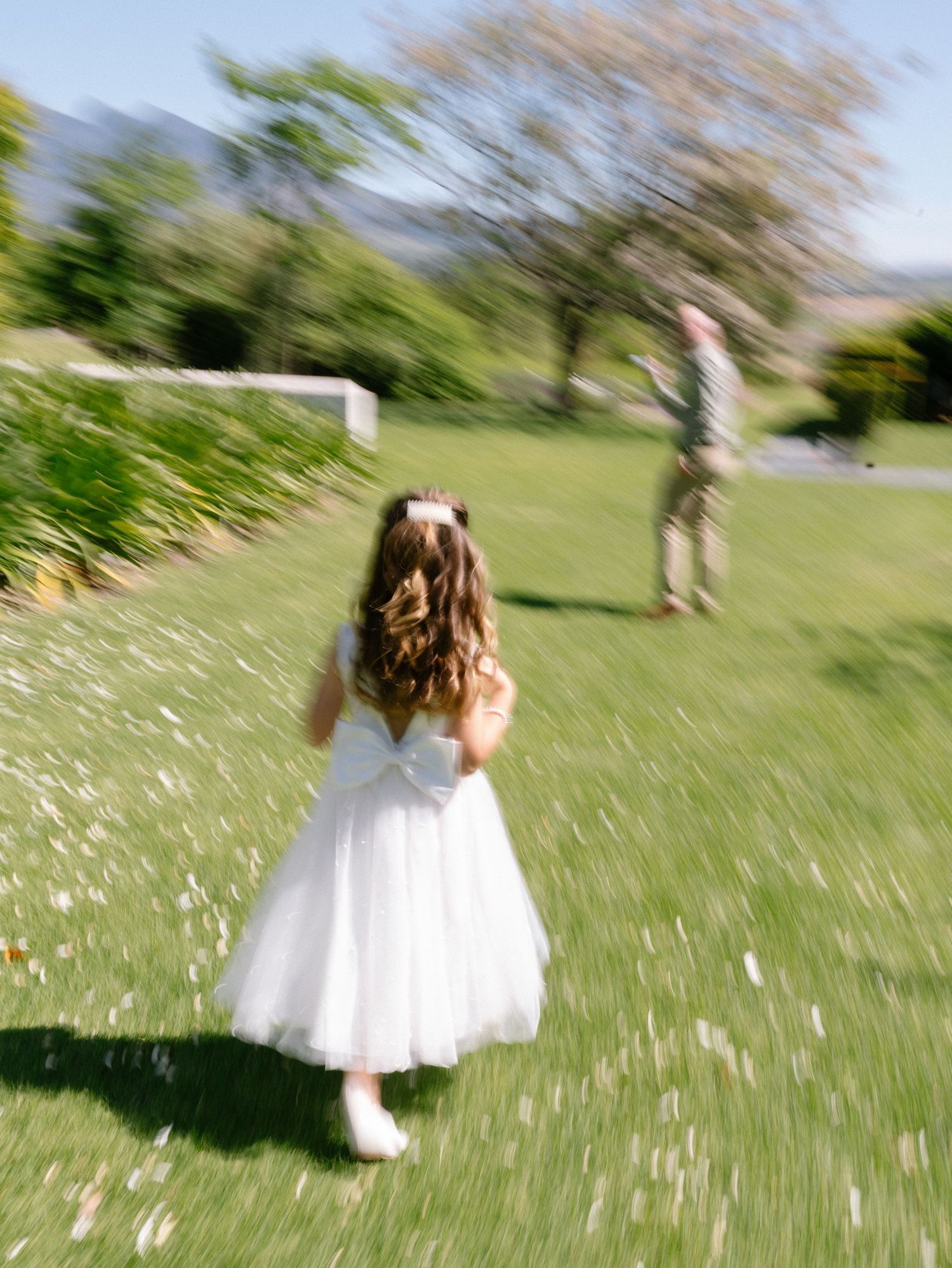 A child in a white tulle dress runs through a sunlit meadow with scattered wildflowers on a spring day.