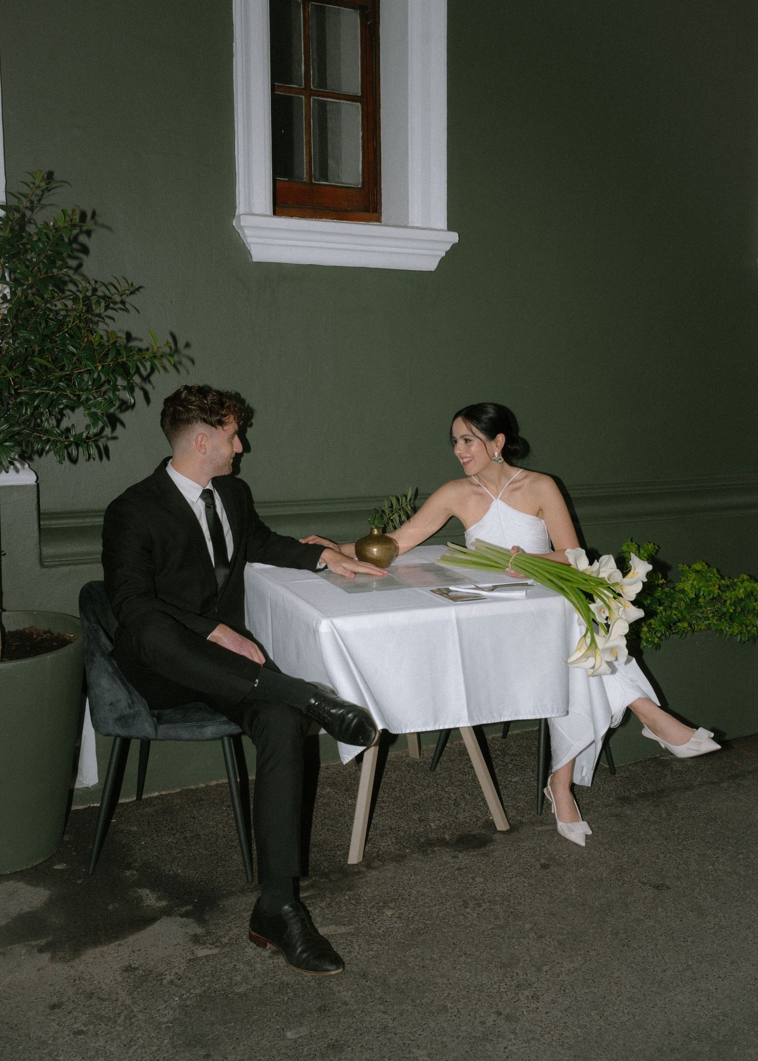 A couple in formal attire sits at a small table with white linens and floral decorations against a green wall at night.