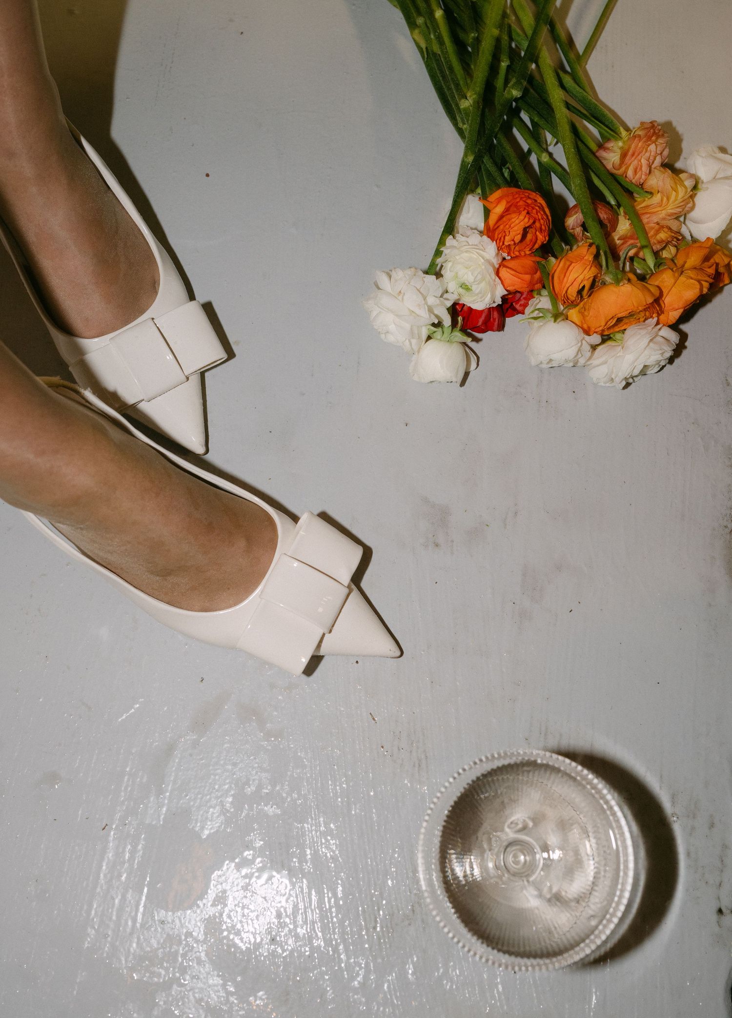 Orange and white flowers lay scattered on a light surface next to beige high heels and a plate.