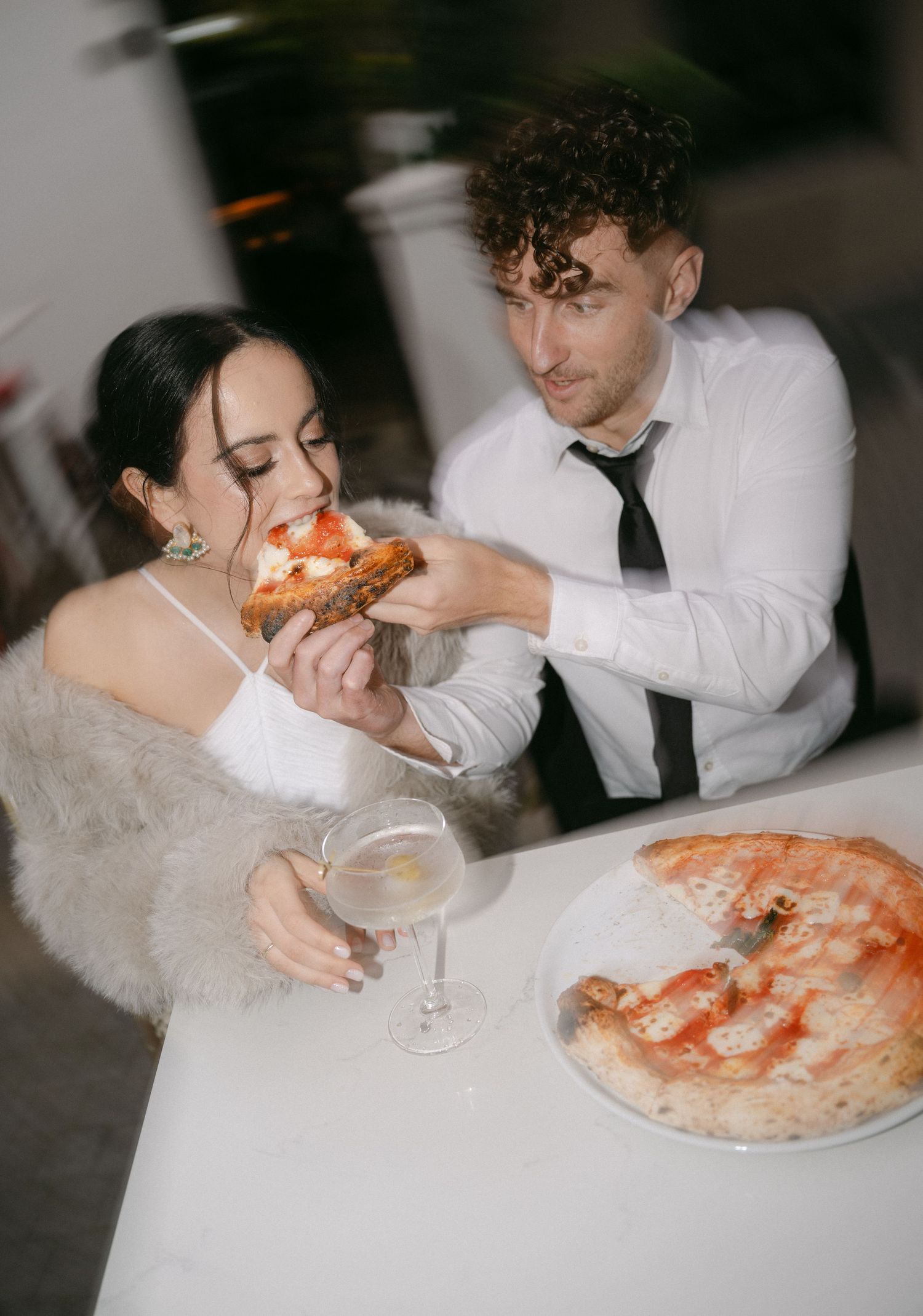 A romantic couple sharing pizza and martinis at a white-clothed table during what appears to be a wedding celebration.