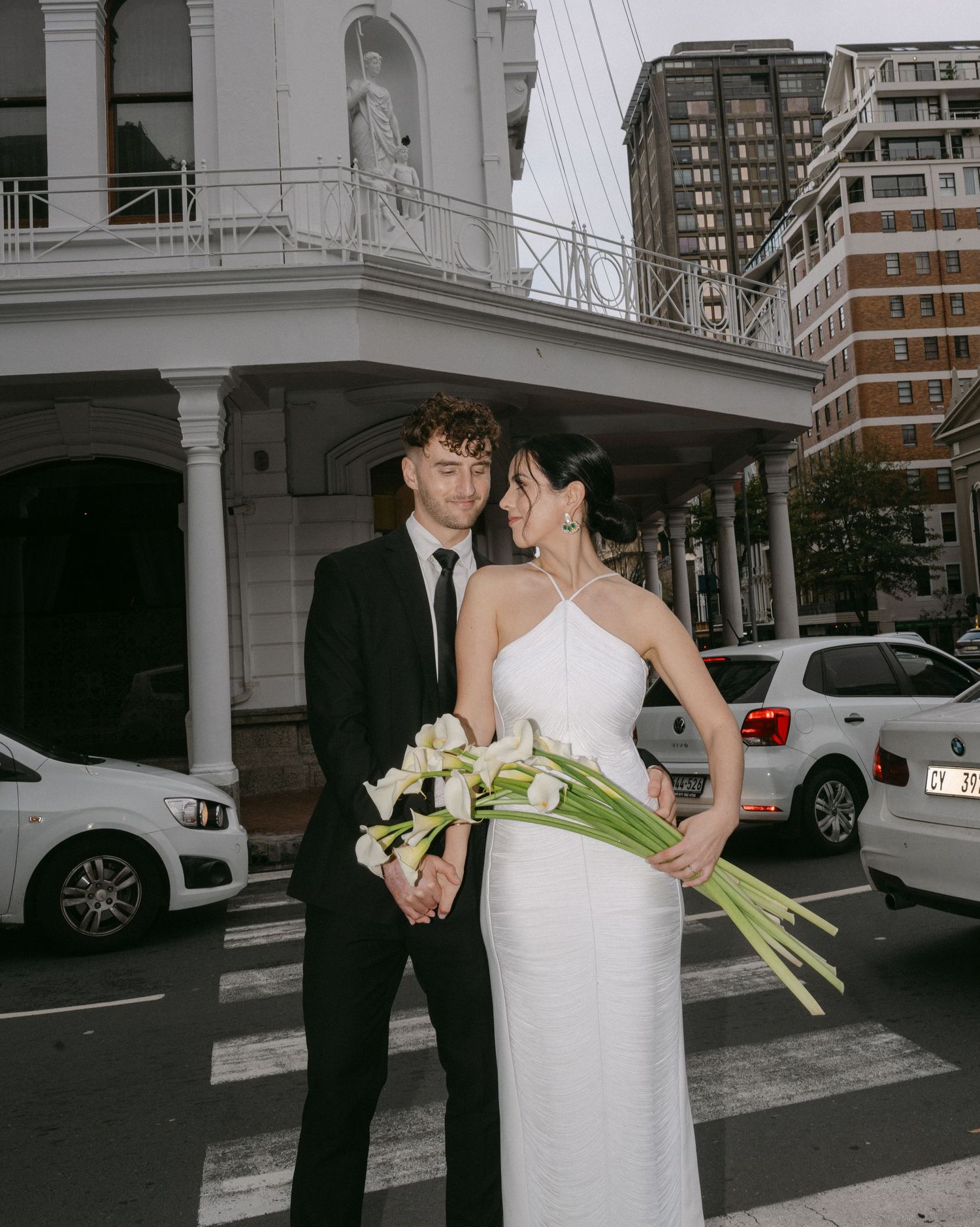 A couple shares an intimate moment outside a modern building with parked cars and urban architecture in the background.