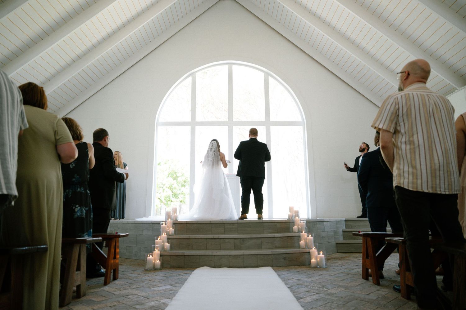 Wedding ceremony taking place in bright white chapel with arched windows and lit candles.