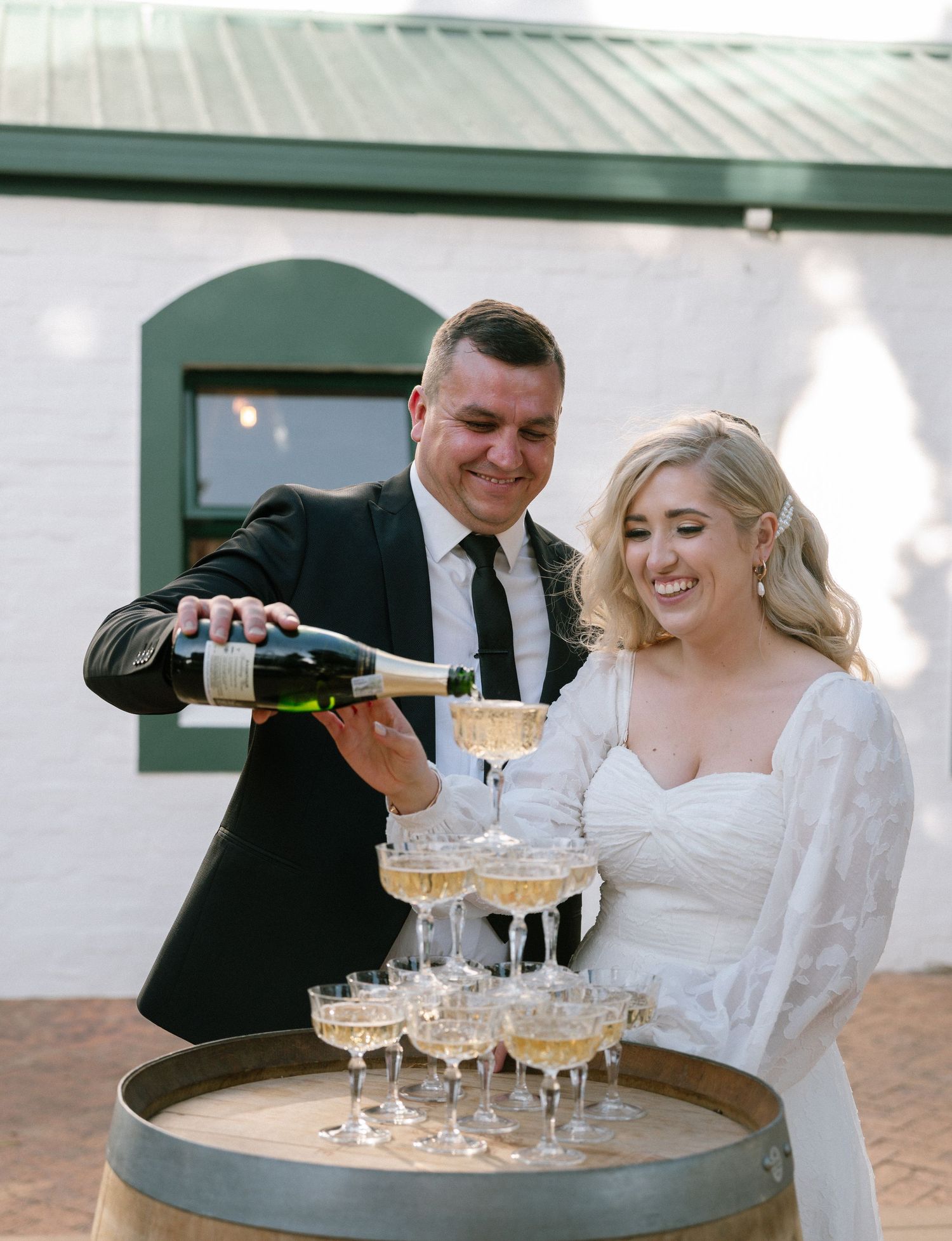 A couple pouring champagne into a tower of glasses at their wedding celebration.