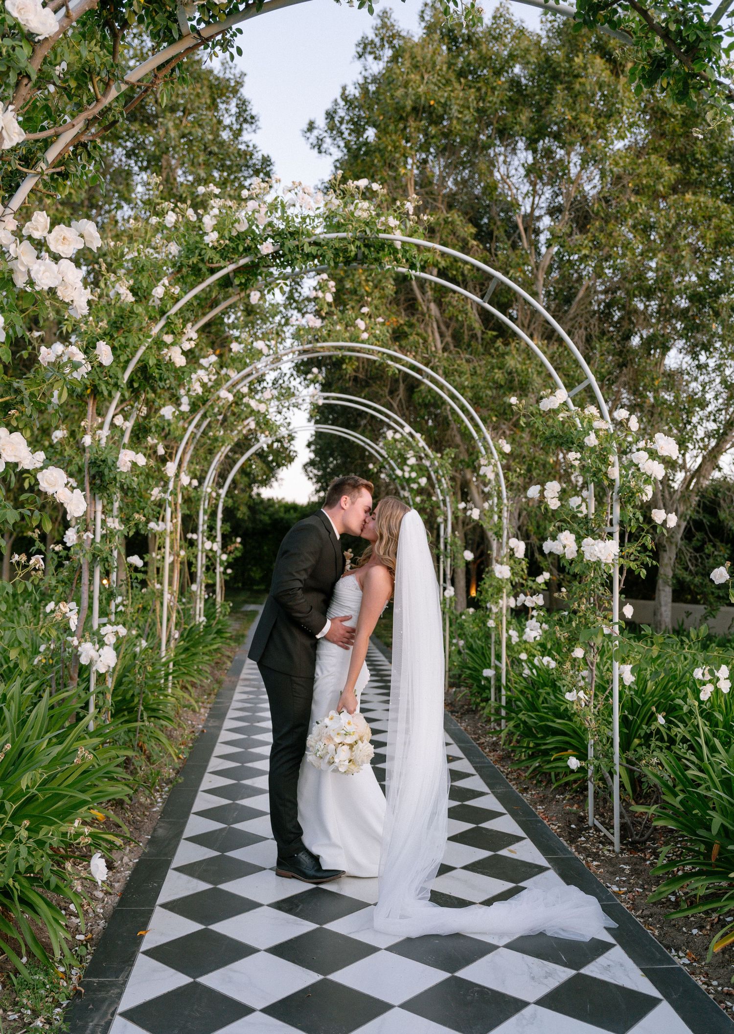 A romantic moment under a floral archway with checkerboard pathway in a garden setting.