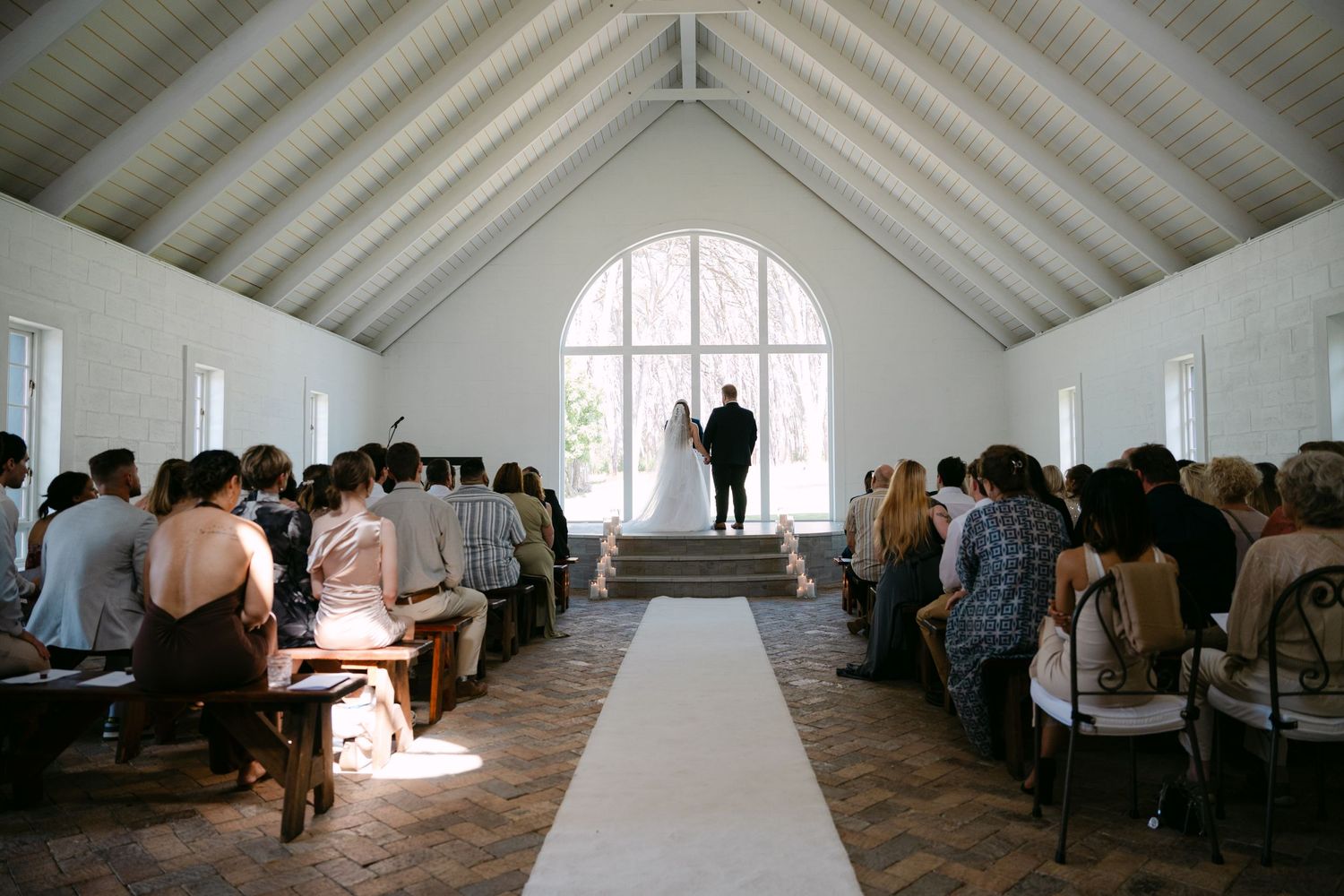 Wedding ceremony in progress inside white chapel with vaulted ceiling and packed pews.