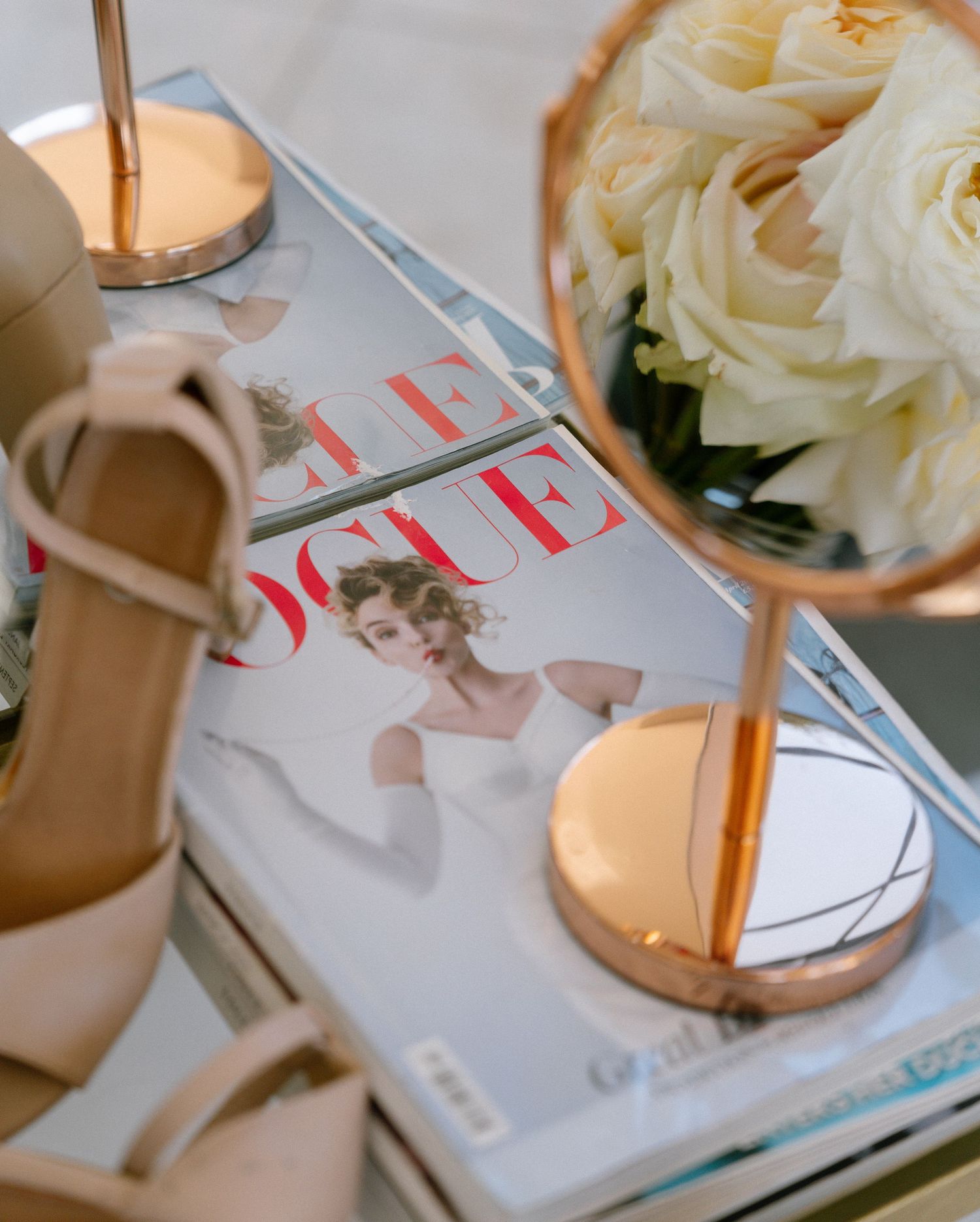 Wedding accessories including beige heels, a fashion magazine, and white roses on a light surface.