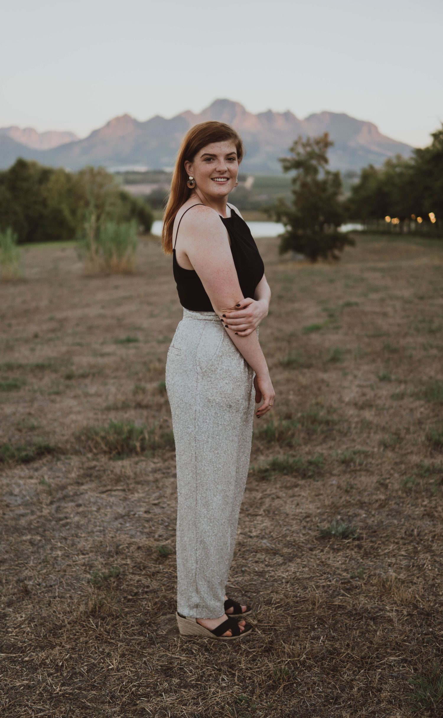 Woman in black top and sparkly pants poses outdoors with mountains in background at sunset.