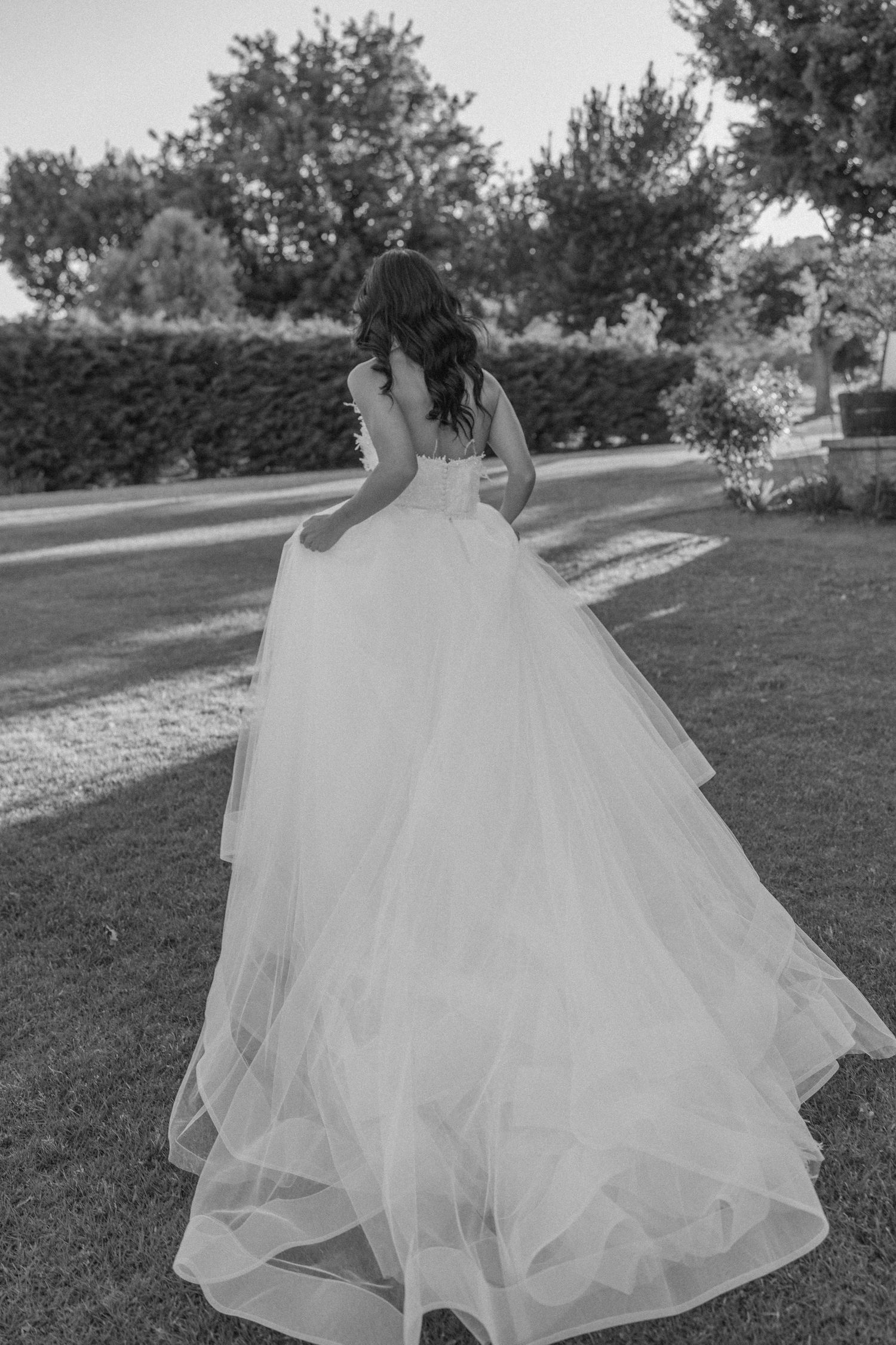 A bride in a flowing tulle ballgown poses against garden landscaping.