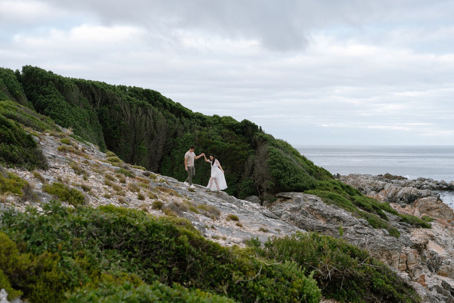 Two figures in white walk along a rocky coastal path overlooking the ocean on a cloudy day.