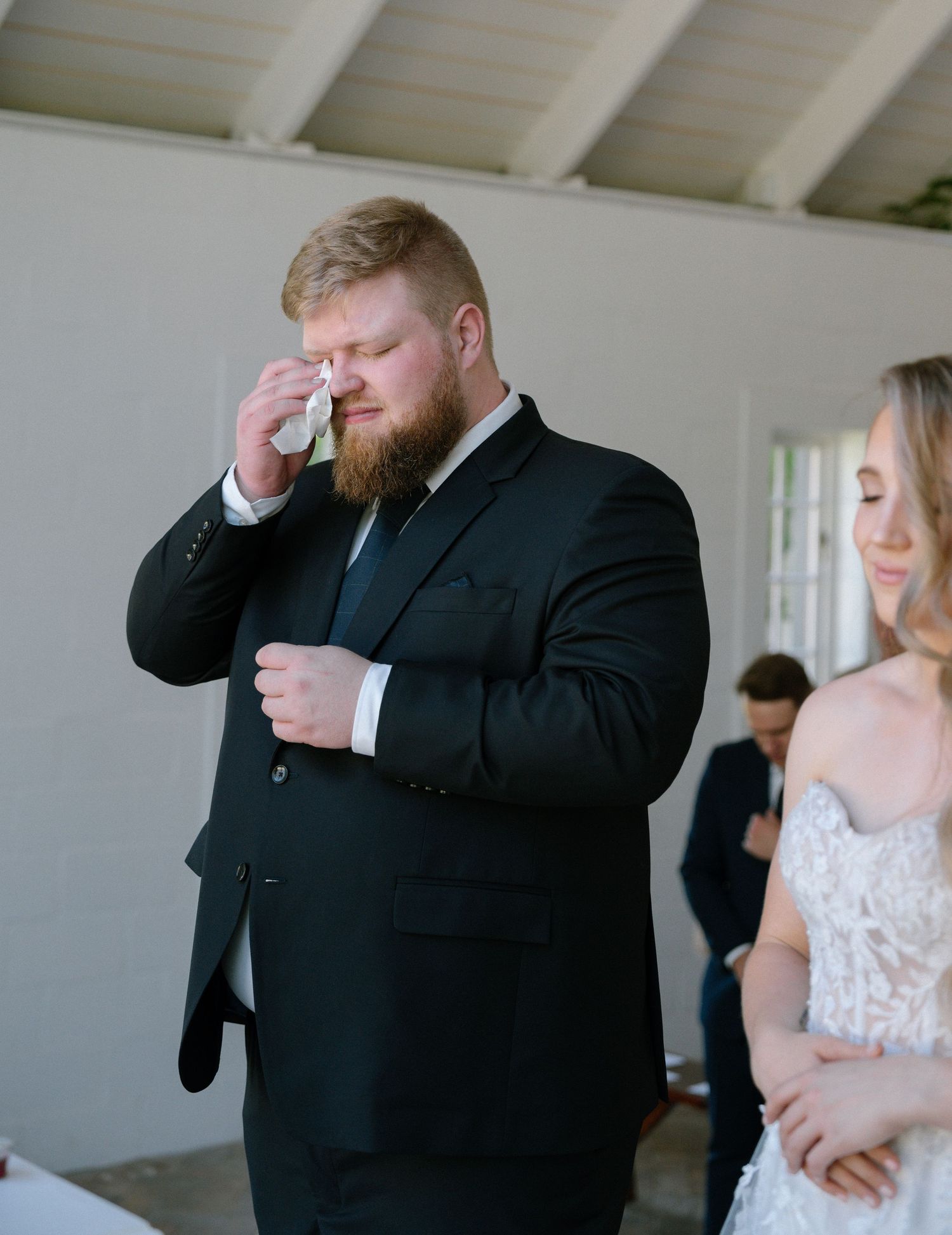 Groom in black suit wipes tears during emotional wedding moment.