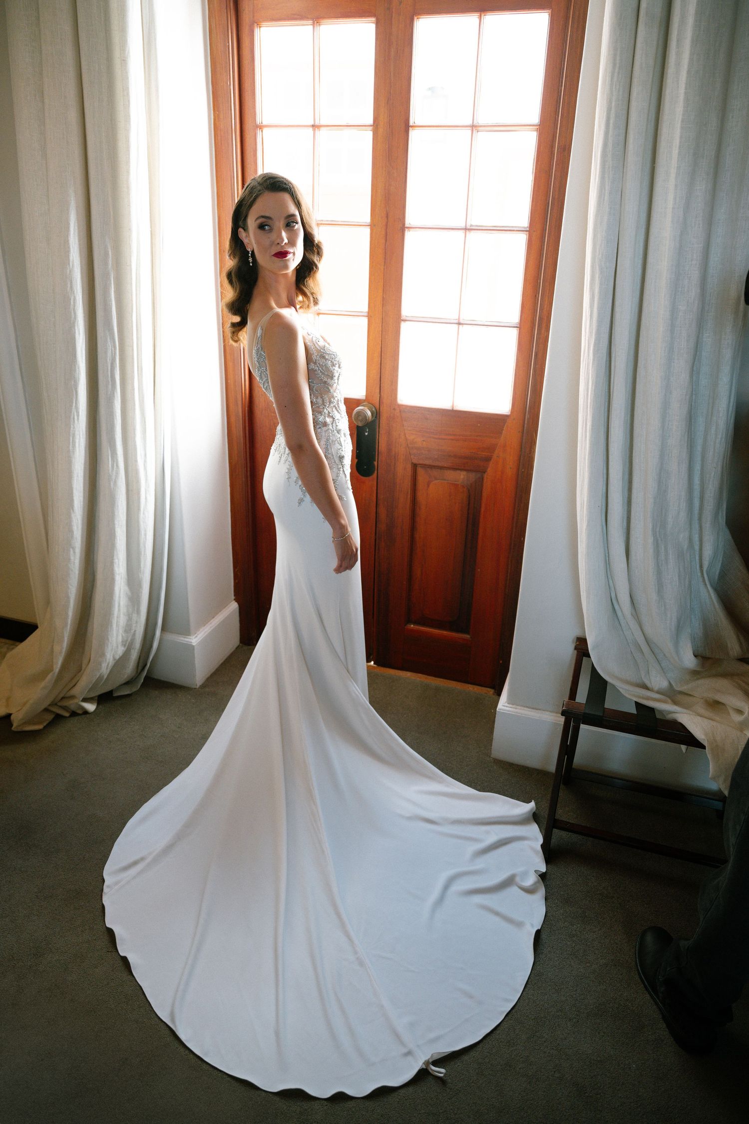 A graceful bride in a fitted white wedding gown with a sweeping train poses elegantly by a wooden door with sheer curtains.