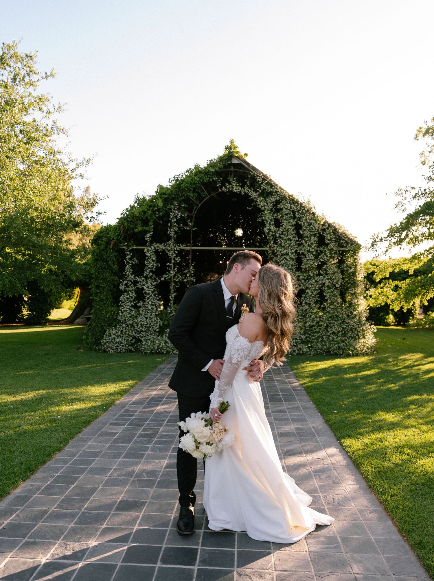 A romantic wedding scene in front of a greenery-covered chapel with stone pathway.