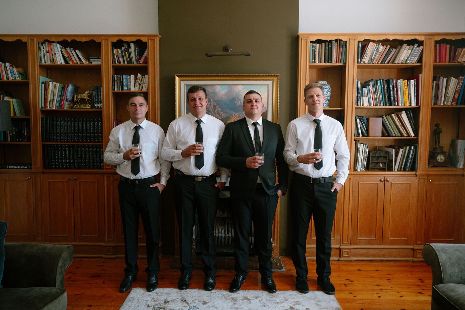 A group stands together in formal attire holding drinks in front of wooden bookshelves in an elegant library setting.