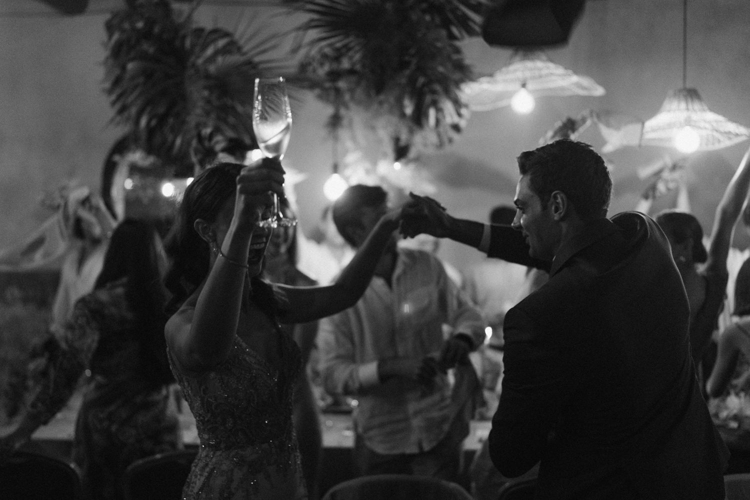 People dance together at a lively party in a black and white photograph with hanging lights and tropical decor.