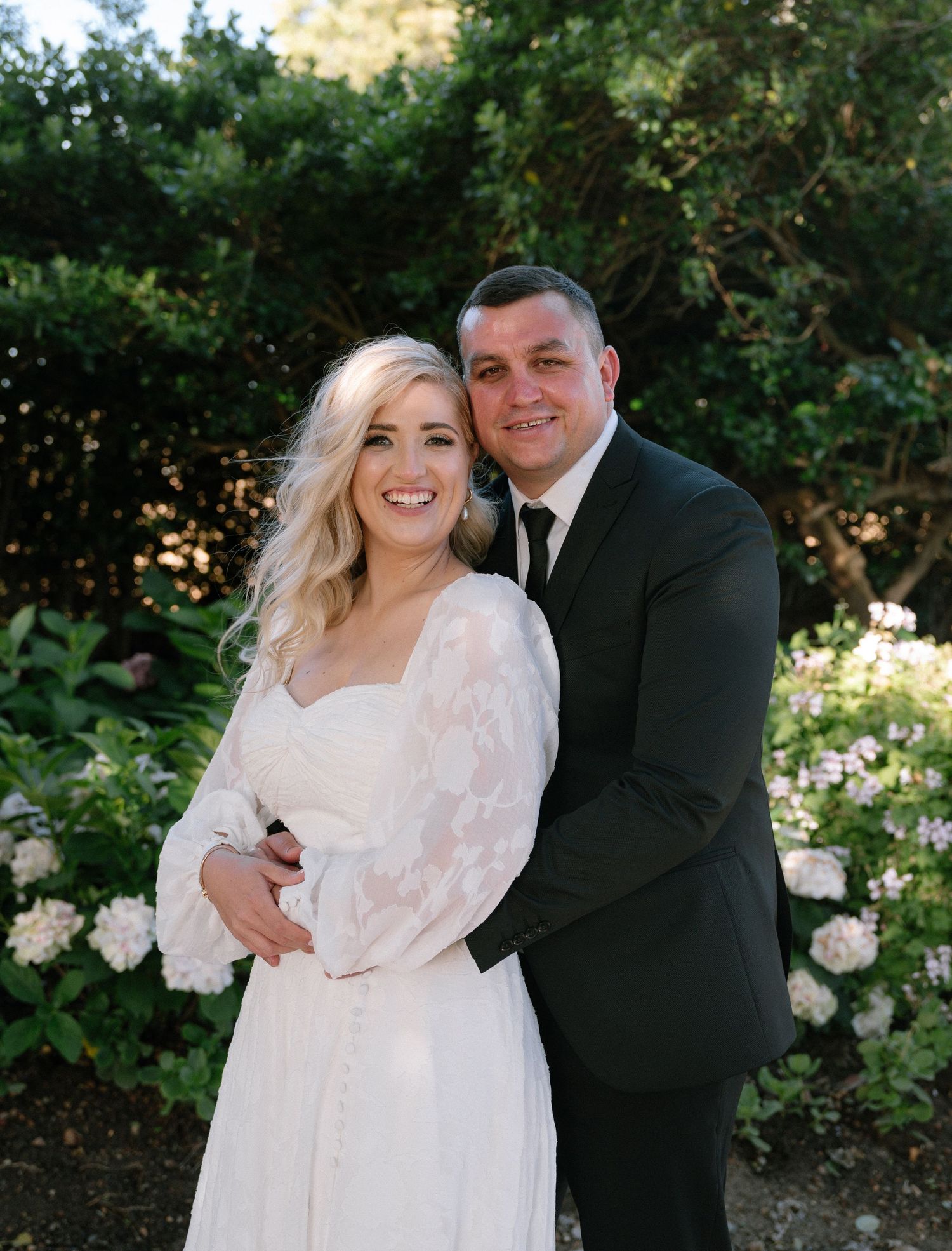A wedding portrait of a couple in formal attire standing in a garden with white flowers.