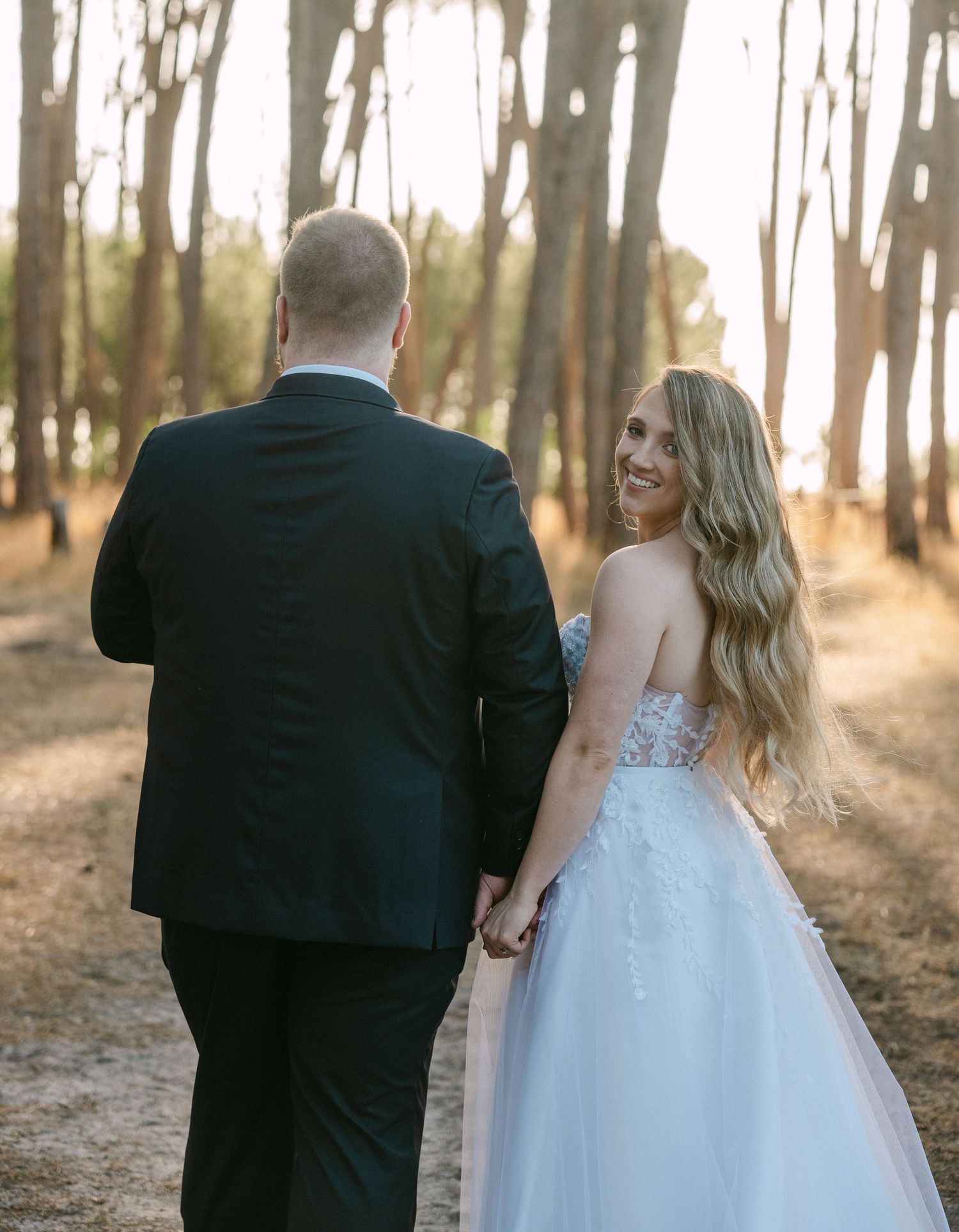 Wedding couple walking through wooded area at sunset, with bride in flowing white gown.