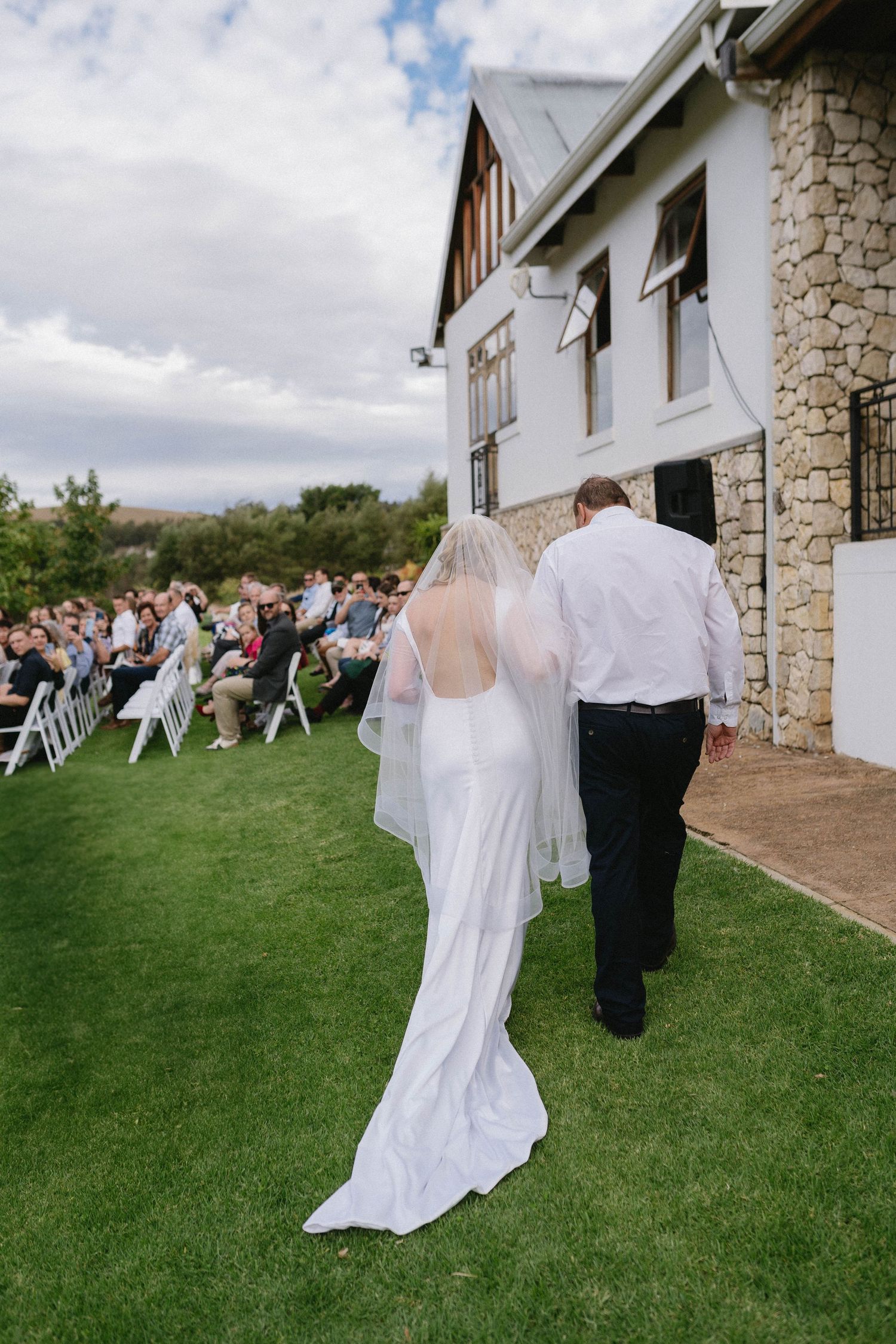 A couple walks on green lawn during outdoor wedding ceremony with guests seated in white chairs by stone building.