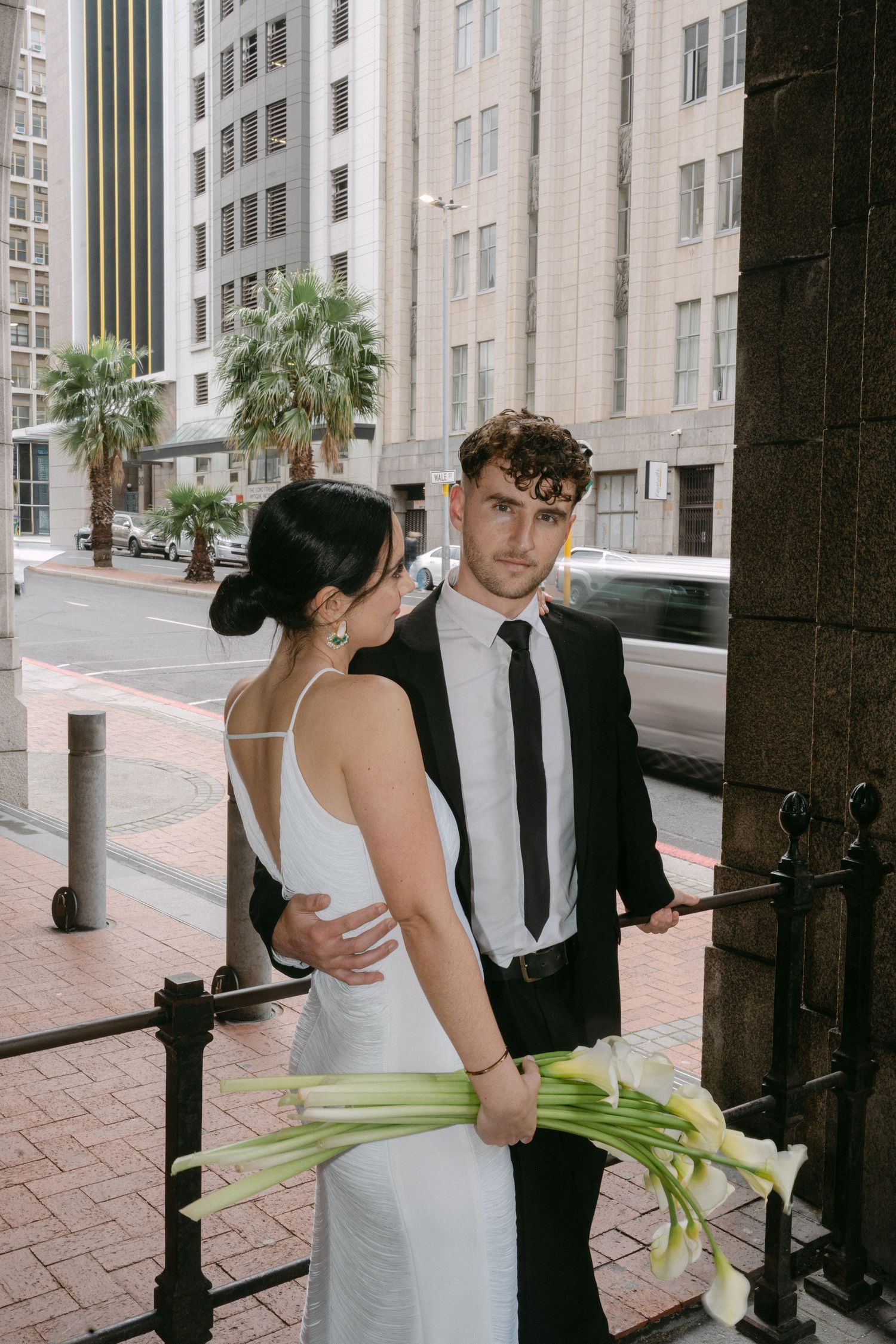 A couple poses against a brick wall with palm trees and modern architecture in the background.